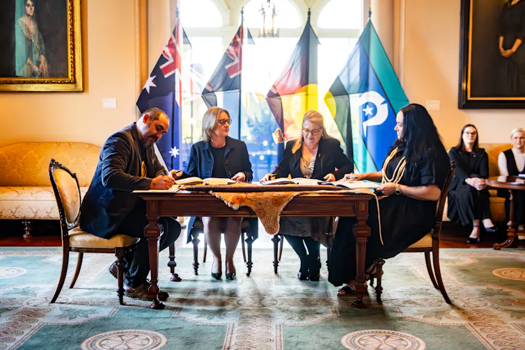 A table with four people signing documents in front of Australian, Aboriginal and Torres Strait Islander flags.