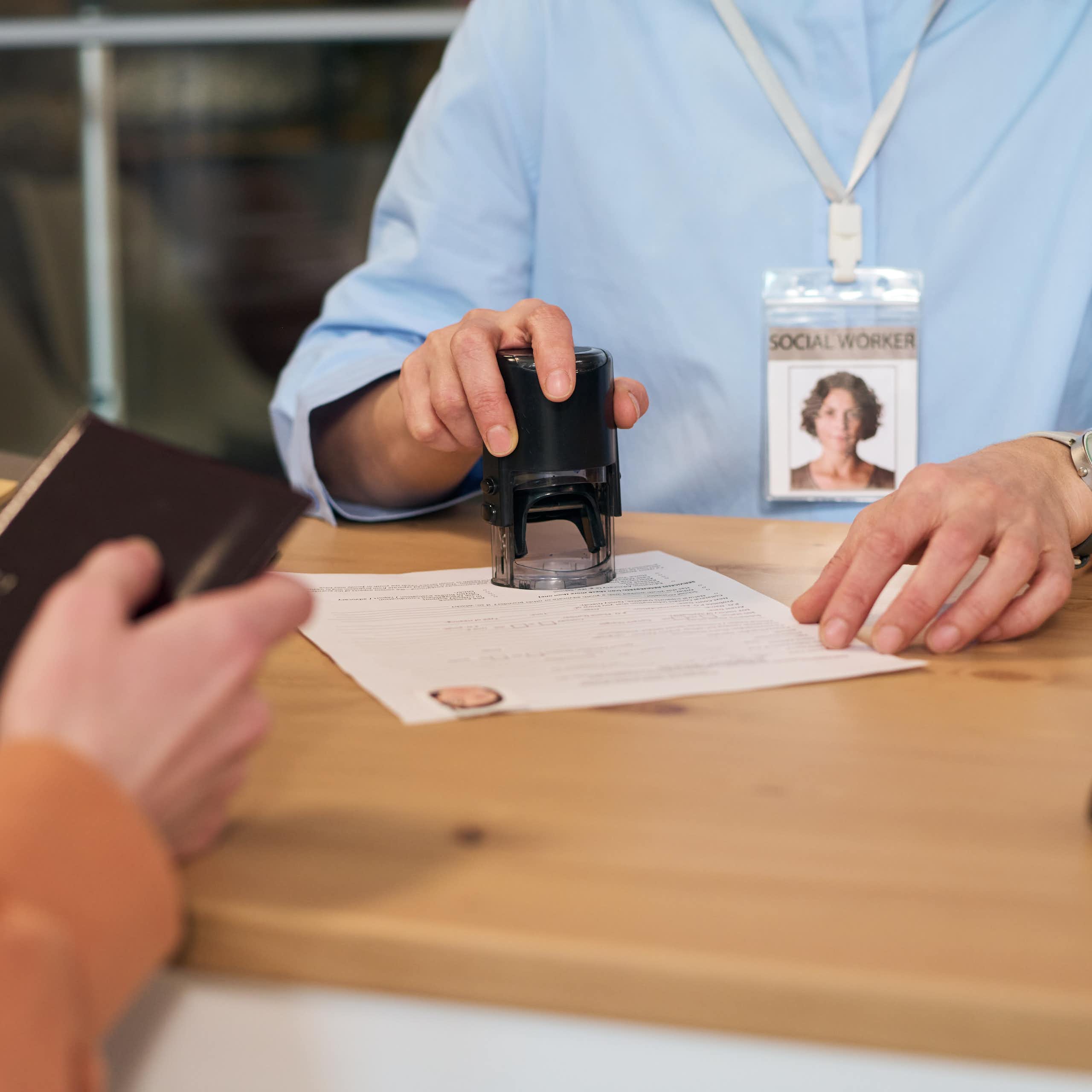 Middle-aged Caucasian social worker with a badge assisting client by stamping an official document in social services office