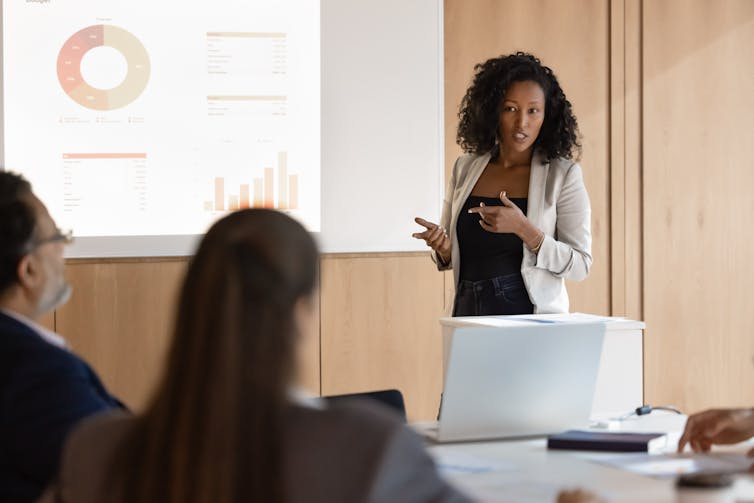 woman delivering a presentation to colleagues with charts and graphs on a screen behind her.