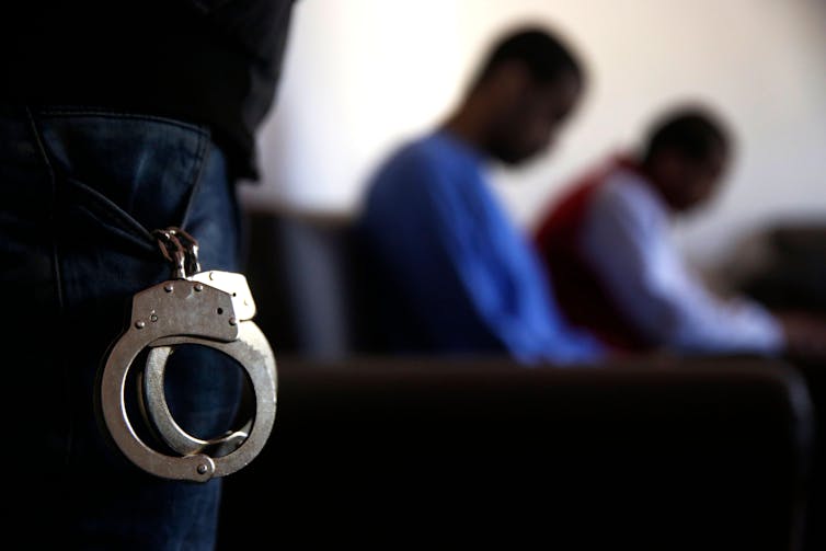 Dois homens negros estão sentados fora de foco no fundo de uma fotografia. NO primeiro plano está um par de algemas.