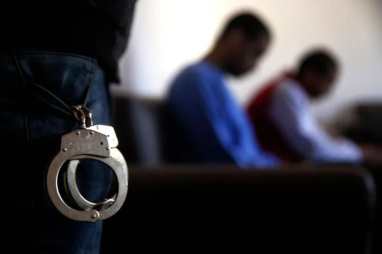 Two black men sit out of focus inthe background of a photograph. IN the foreground is a pair of handcuffs.
