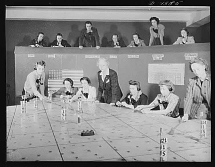 Las mujeres han estado cartografiando el mundo durante siglos y ahora defienden a las personas que han quedado fuera de esos mapas. 1 Fotografía en blanco y negro de mujeres rodeando una mesa de celosía, una persona empujando piezas a través de las superficies mientras otras miran al balcón.