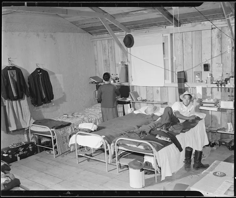 People stand and sit near beds in an open space with clothes hanging from hooks on the wooden wall.