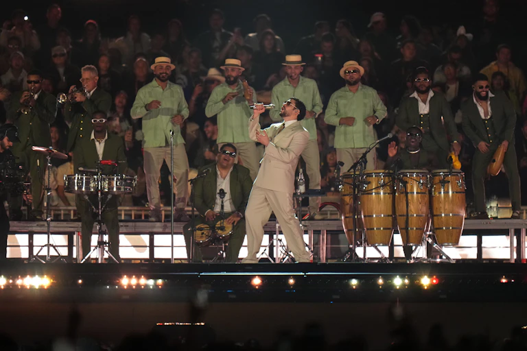 A Puerto Rican man in a beige suit and sunglasses sings into a microphone in stage