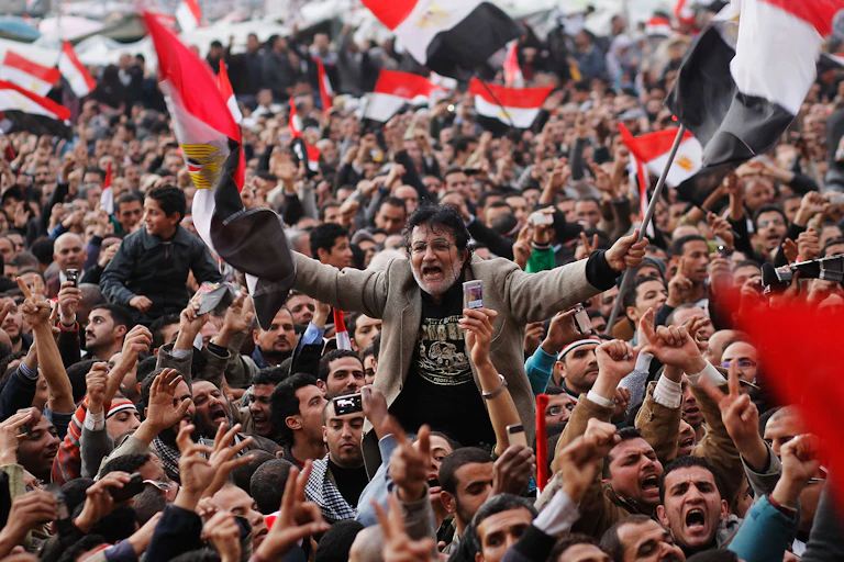 A protester is carried on shoulders through a crowded square as people wave flags with red, white and black colors.