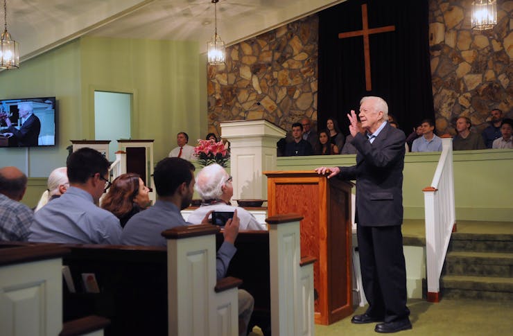 An elderly man in a dark suit stands in front of a church congregation, raising a hand in greeting.