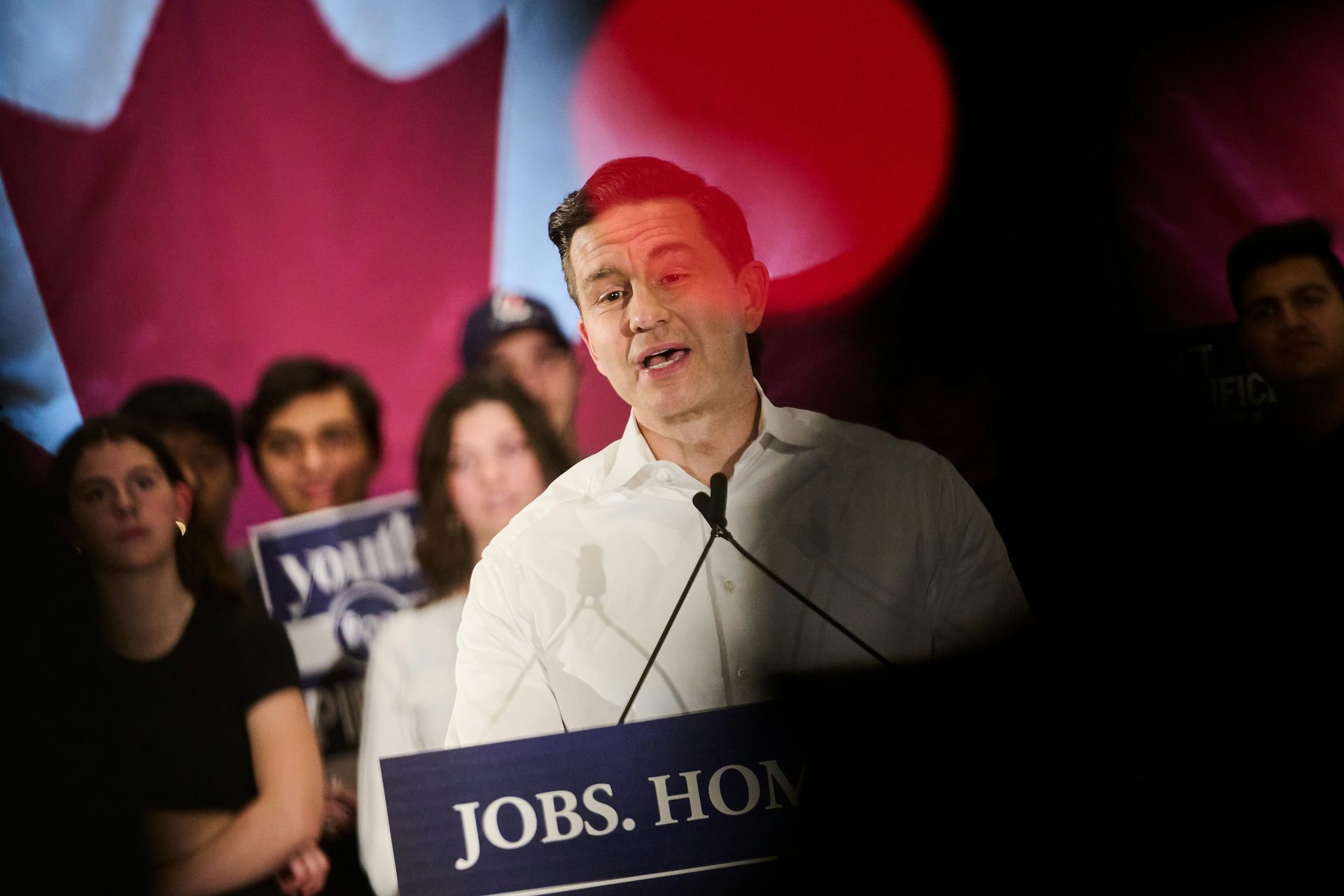 A man with dark hair speaks into a microphone as young people stand behind him.