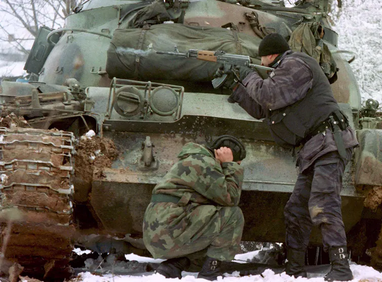 A man in military camouflage crouches while covering his ear next to an army tank.