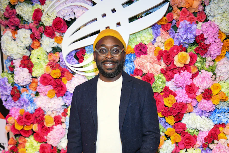 A man dressed in dress jacket, jeans and yellow beanie stands on a red carpet with a floral display behind him.