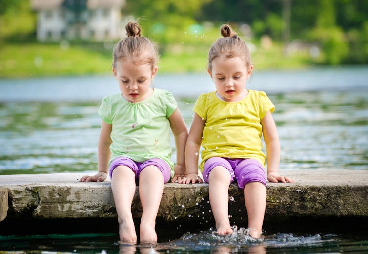 Identical female twins sitting on a wall, their feet in the water.