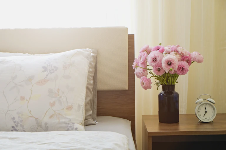 Small, pretty vase of flowers and analogue alarm clock on the bedside next to pillows.