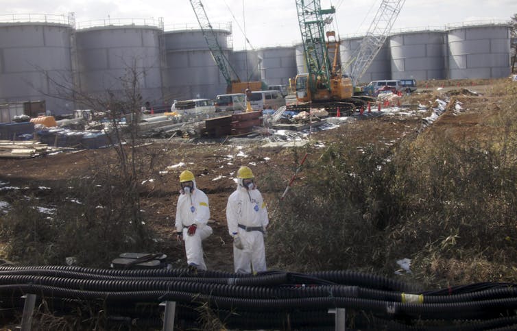 Two people in white hazmat suits among power plant ruins.