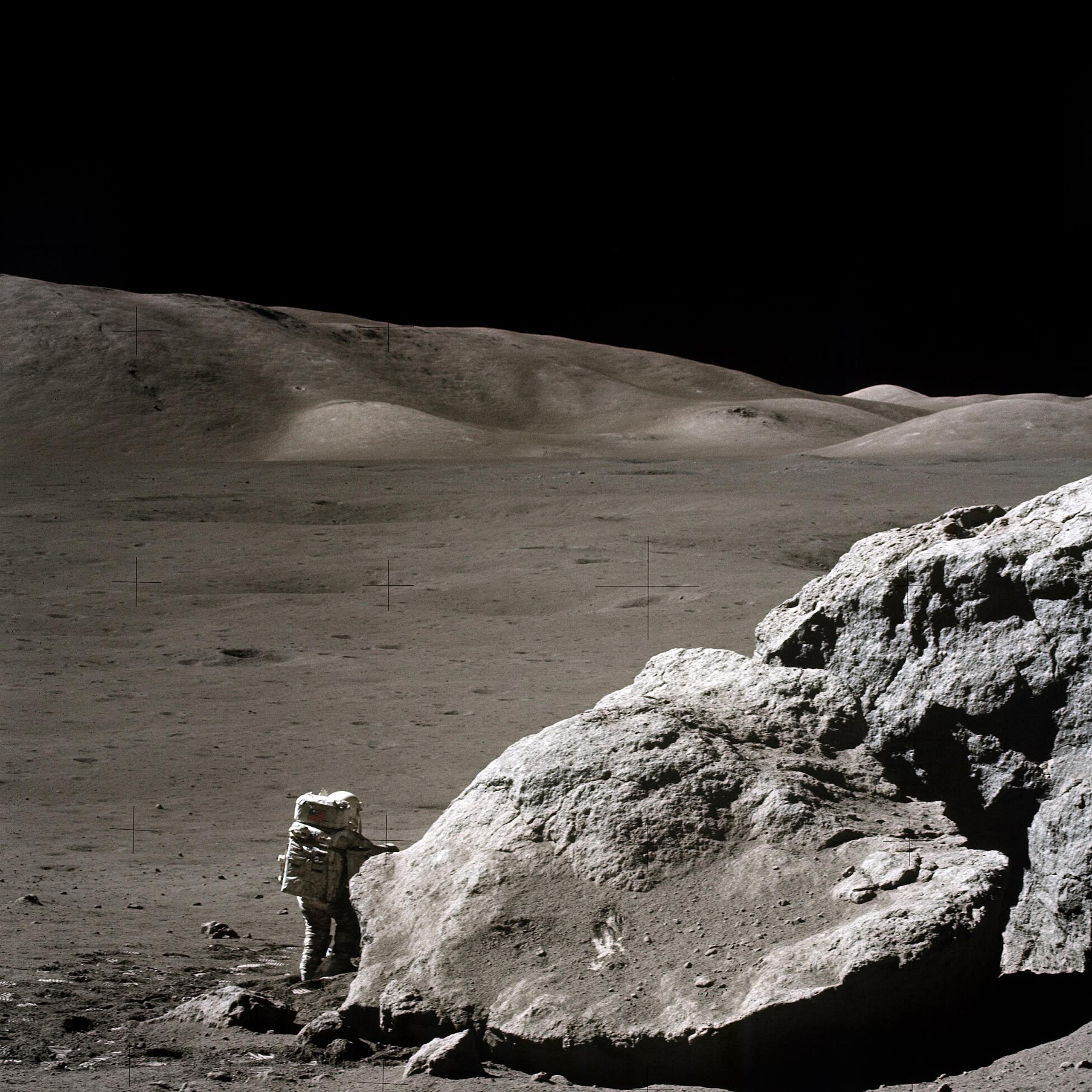 Photographie de la surface de la Lune. Un astronaute en combinaison se tient près d'un grand rocher. De l'autre côté, un véhicule est stationné.