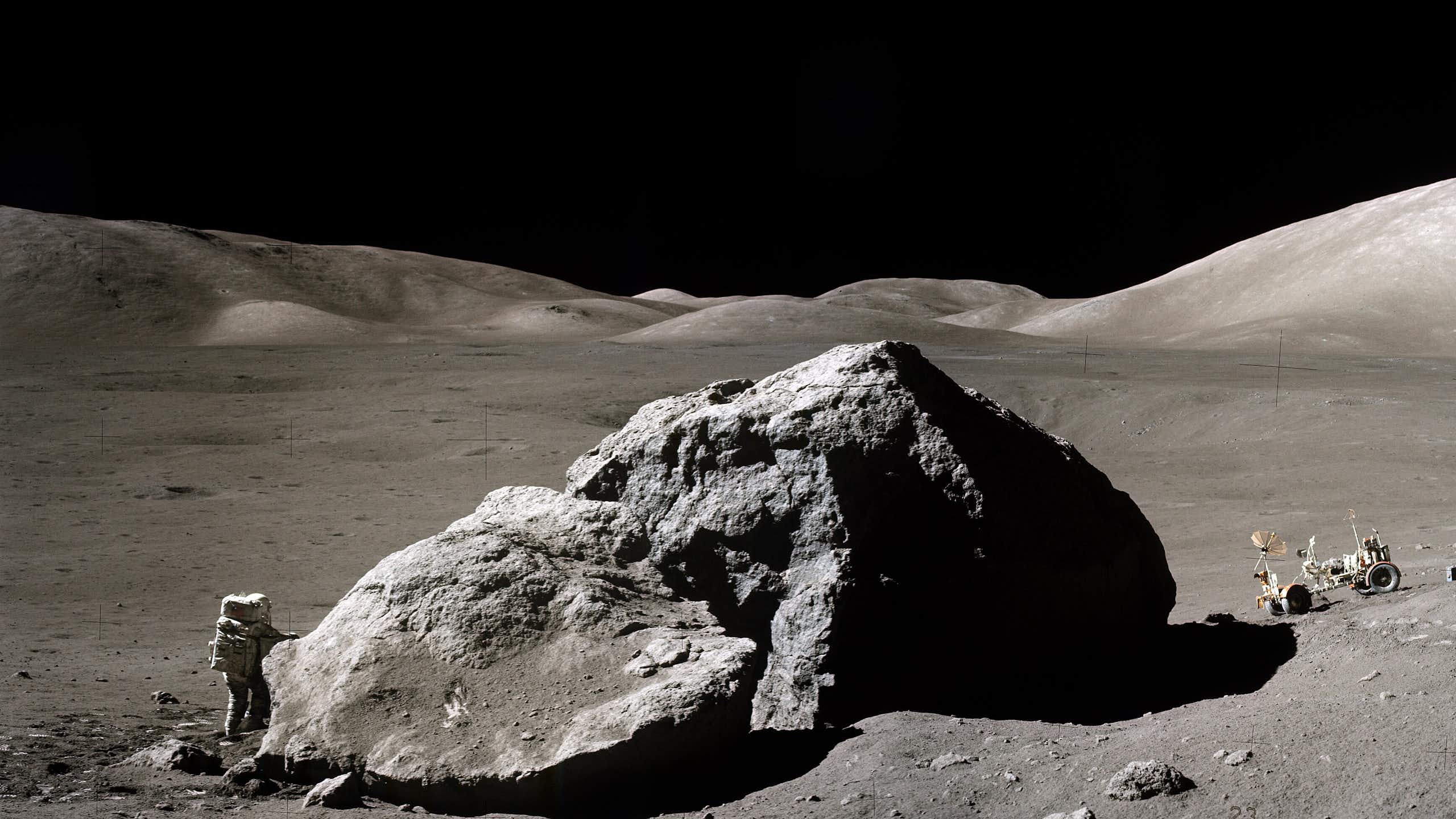 Photographie de la surface de la Lune. Un astronaute en combinaison se tient près d'un grand rocher. De l'autre côté, un véhicule est stationné.