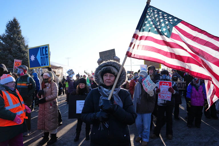 Protesters march down the street in Minneapolis, led by a woman holding a large U.S. flag.