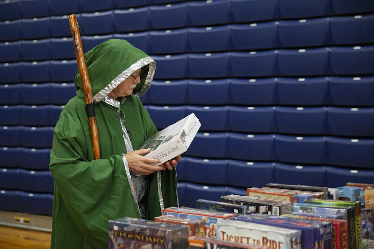 A man wearing a green hooded robes and carrying a wooden staff holds a boxed board game.
