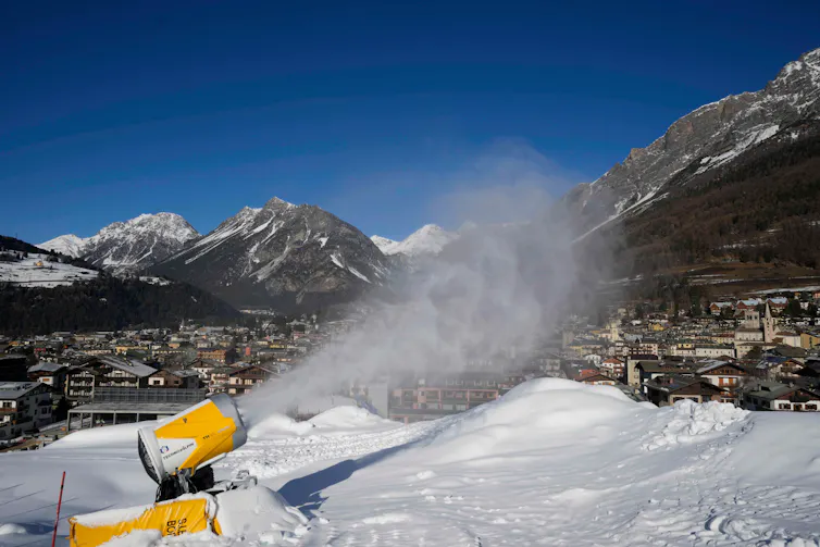 A cylindrical device sprays snow over an area, mountains are seen in the background