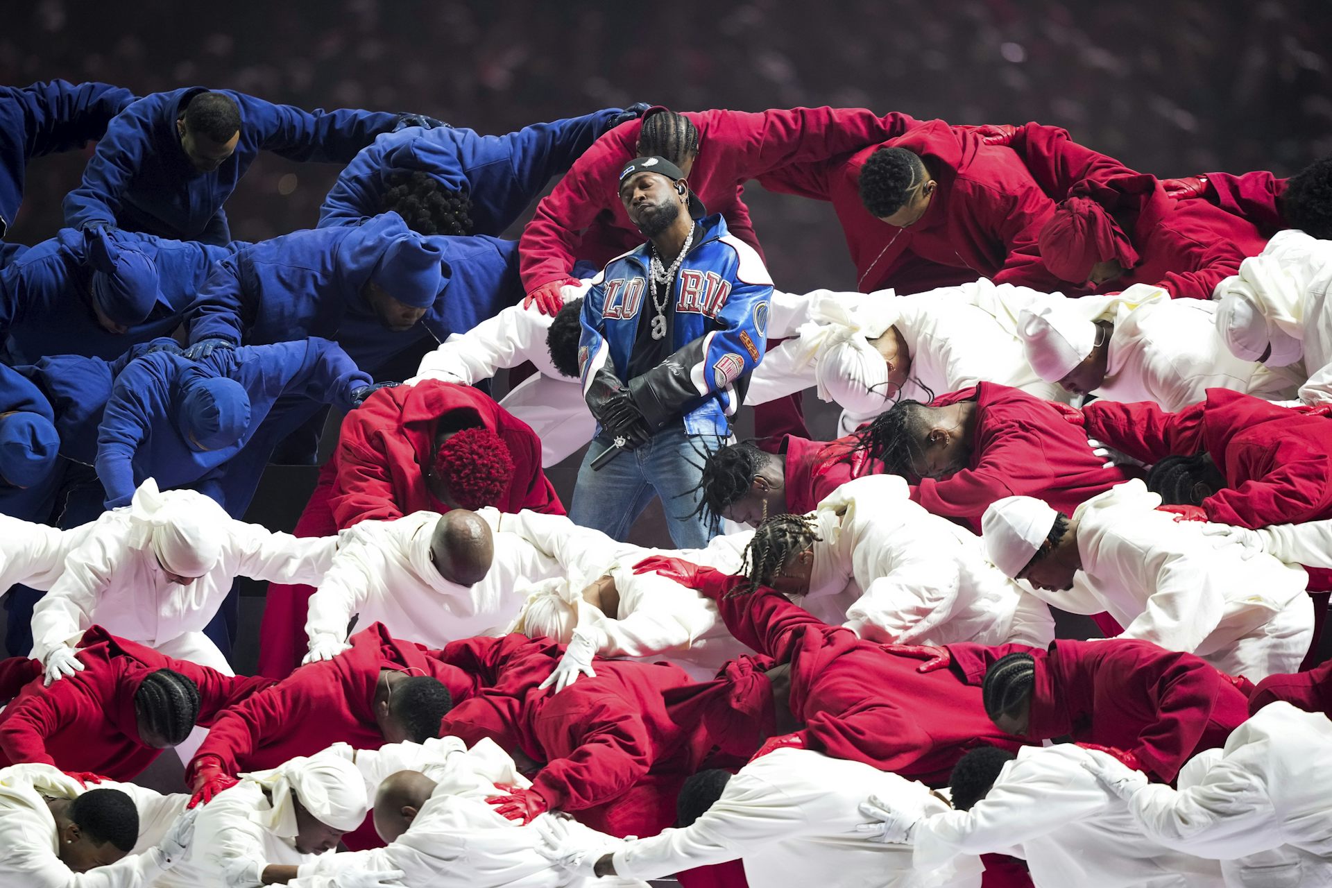 Un hombre negro con una chaqueta de cuero azul y blanca con Gloria en el frente se encuentra en medio de un grupo de bailarines vestidos de rojo, blanco y azul.