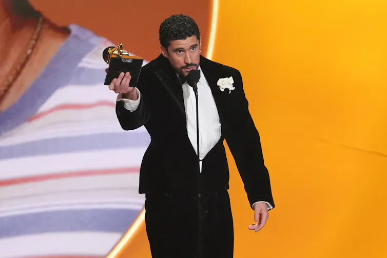 A Puerto Rican man in a black tuxedo speaks into a microphone while holding a Grammy trophy