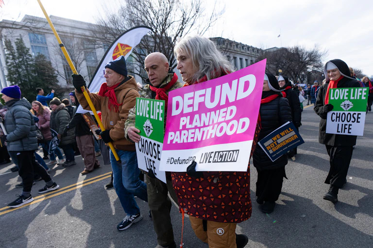 Anti-abortion protesters, one carrying a sign saying "Defund Planned Parenthood," march in a cluster.