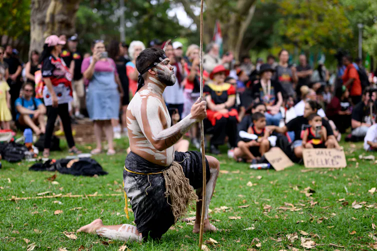 An Aboriginal man in ceremonial dress kneels on one knee on the grass