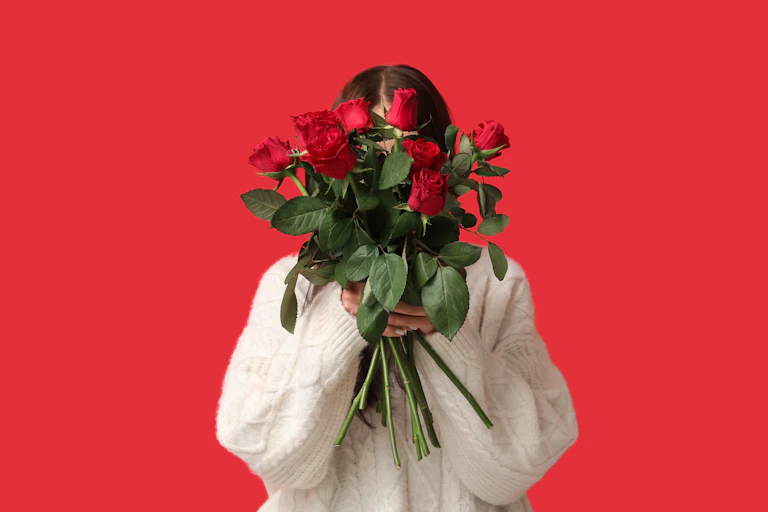 Woman against red background holding bunch of red roses in front of her face.