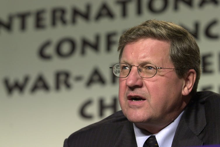 A man with light brown hair and glasses speaks in front of a banner that reads International Conference on War-Affected Children
