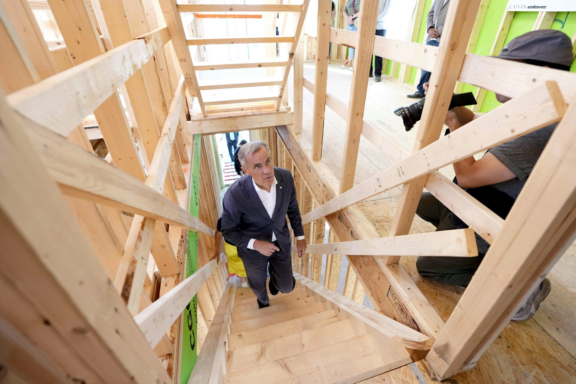 A slender man with short grey hair walks up a wooden staircase in a house under construction.