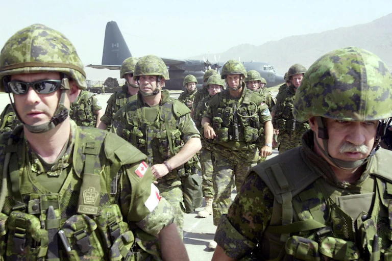 Men in camouflage and wearing helmets walk en masse from a military transport plane on a tarmac.