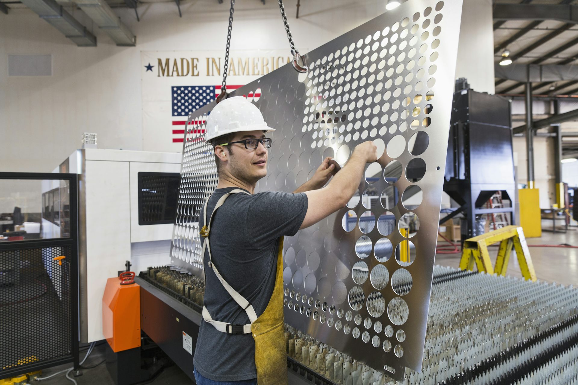 A man wearing a white safety helmet holds up a piece of sheet metal with holes punched through it.