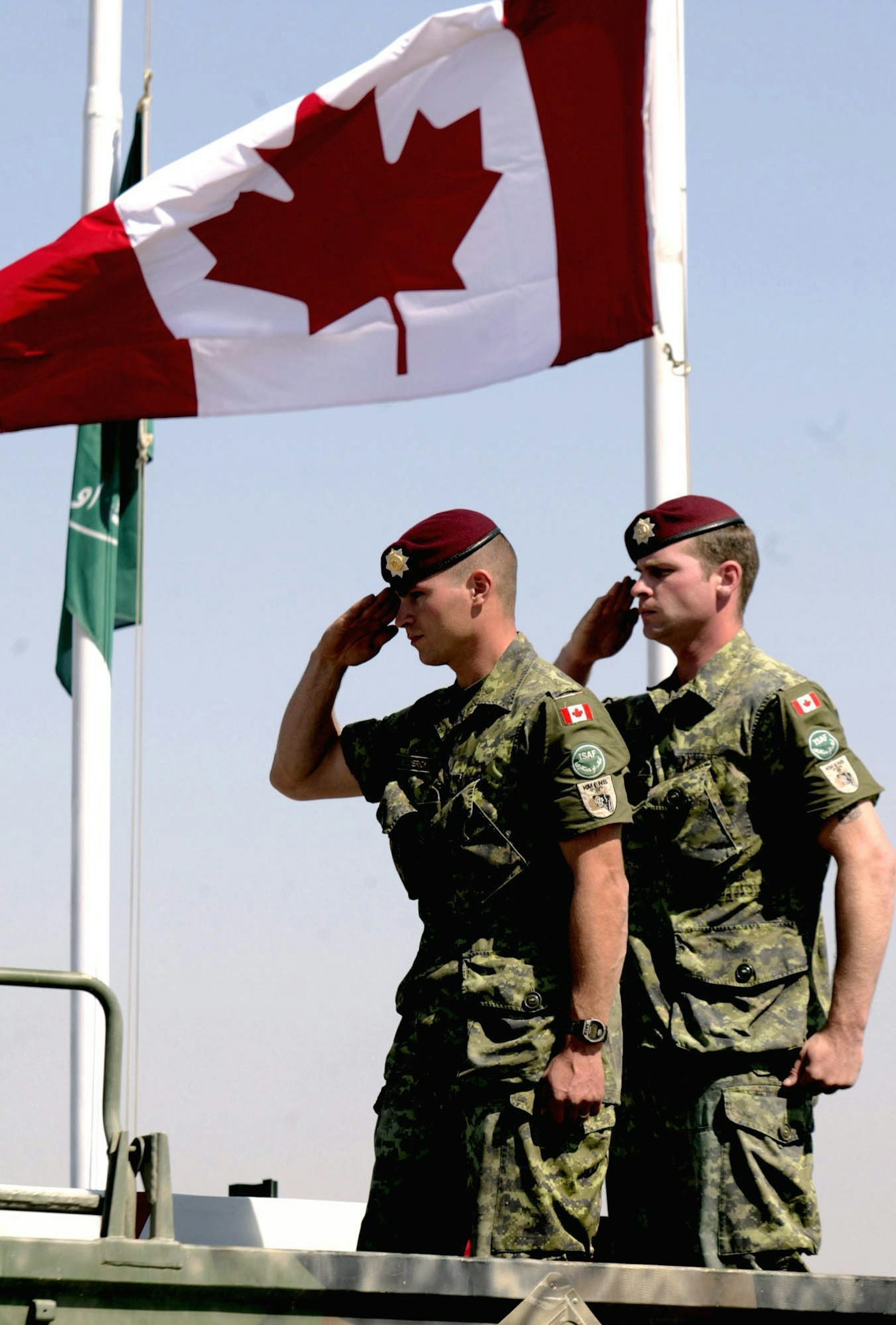 Two Canadian soldiers salute as a Canadian flag flies above them.
