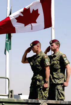 Two Canadian soldiers salute as a Canadian flag flies above them.