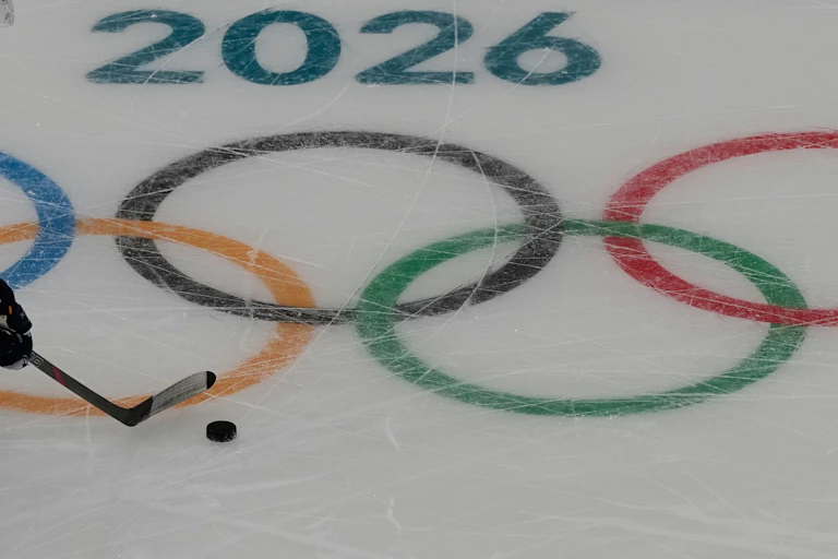 An ice rink with '2026' and the olympic rings drawn on it, and a hockey player pushing a puck across it.