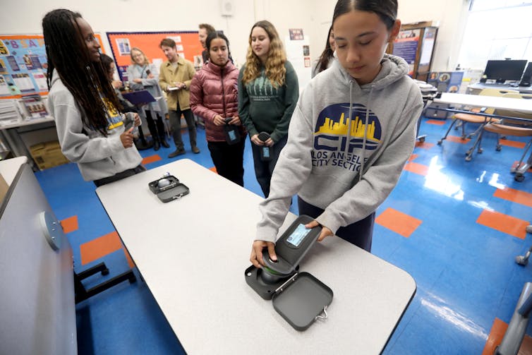 A teenage girl stands at a table holding a pouch near a group of other young people.