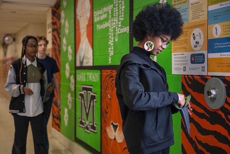 Colleges are more and more telling scholars they should put their telephones away – Ohio’s instance presentations combined effects following new bans 1 A young girl, as seen from the side, looks down at a dark pouch in a colorful hallway.