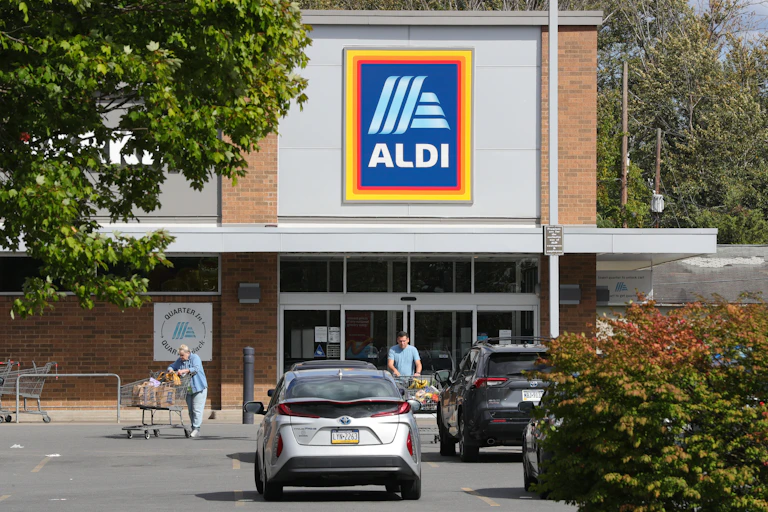 A store front with the text " Aldi" is pictured with shoppers and cars in front.