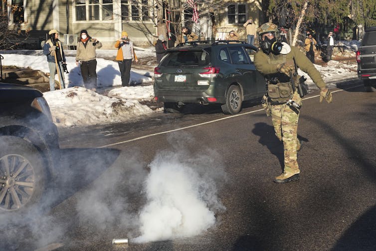 An agent dressed in tactical gear throws a canister emitting smoke on the road. In the background stand onlookers holding cameras.