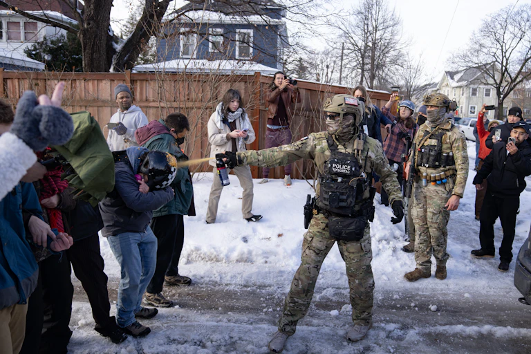 An agent dressed in tactical gear sprays pepper spray directly at a ducking protestor's head from about a foot away.