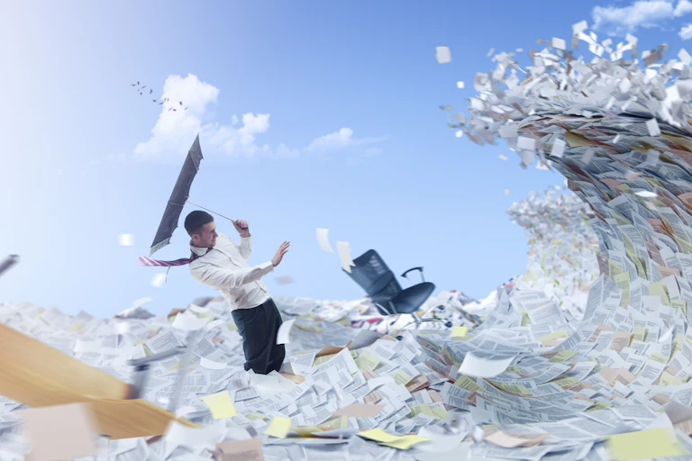 a man carrying an inverted umbrella leans away as a tidalwave made of paper documents heads toward him