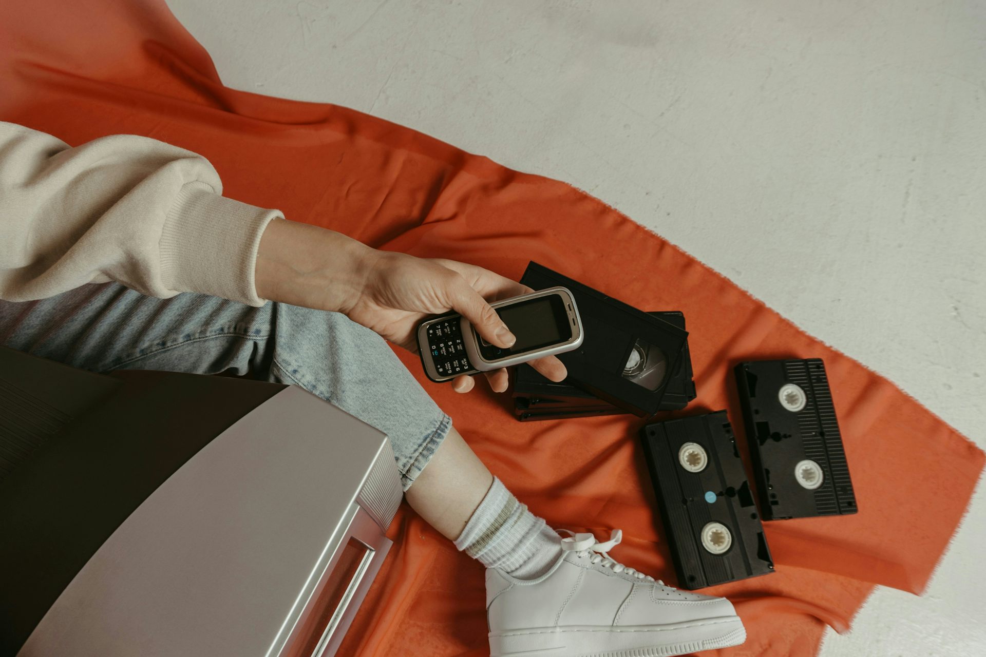 A person holding a flip phone sitting on the ground beside VHS tapes