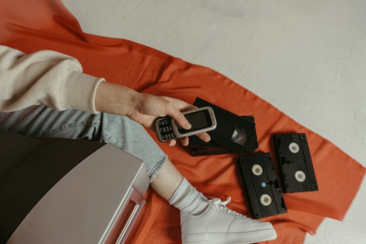 A person holding a flip phone sitting on the ground beside VHS tapes