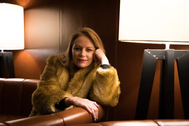 A blonde woman in a fur sits on a leather couch.