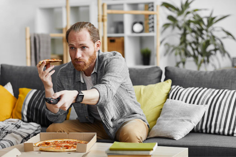 A young man sitting on a couch in a stylish apartment, eating pizza and watching television