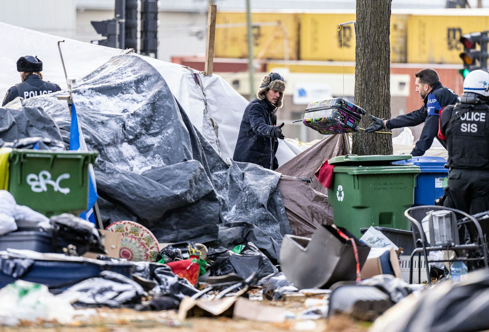 A police officer tosses an unhoused man a suitcase in an encampment.