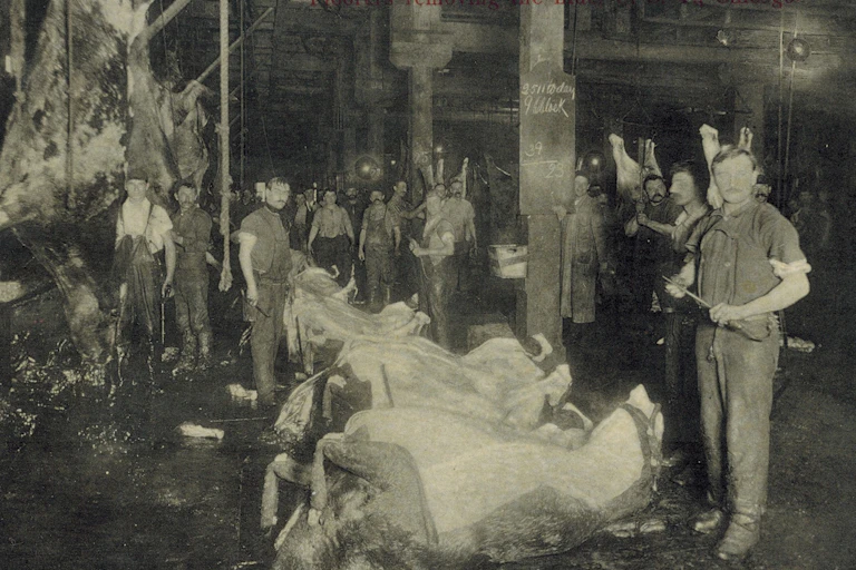 Men stand holding tools in front of beef caracasses.