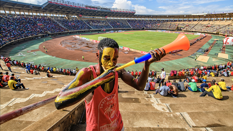 Um homem pintado de amarelo, preto e vermelho com coletes vermelhos segura uma buzina de plástico e sopra nela, em pé no topo dos assentos de um grande estádio de futebol.