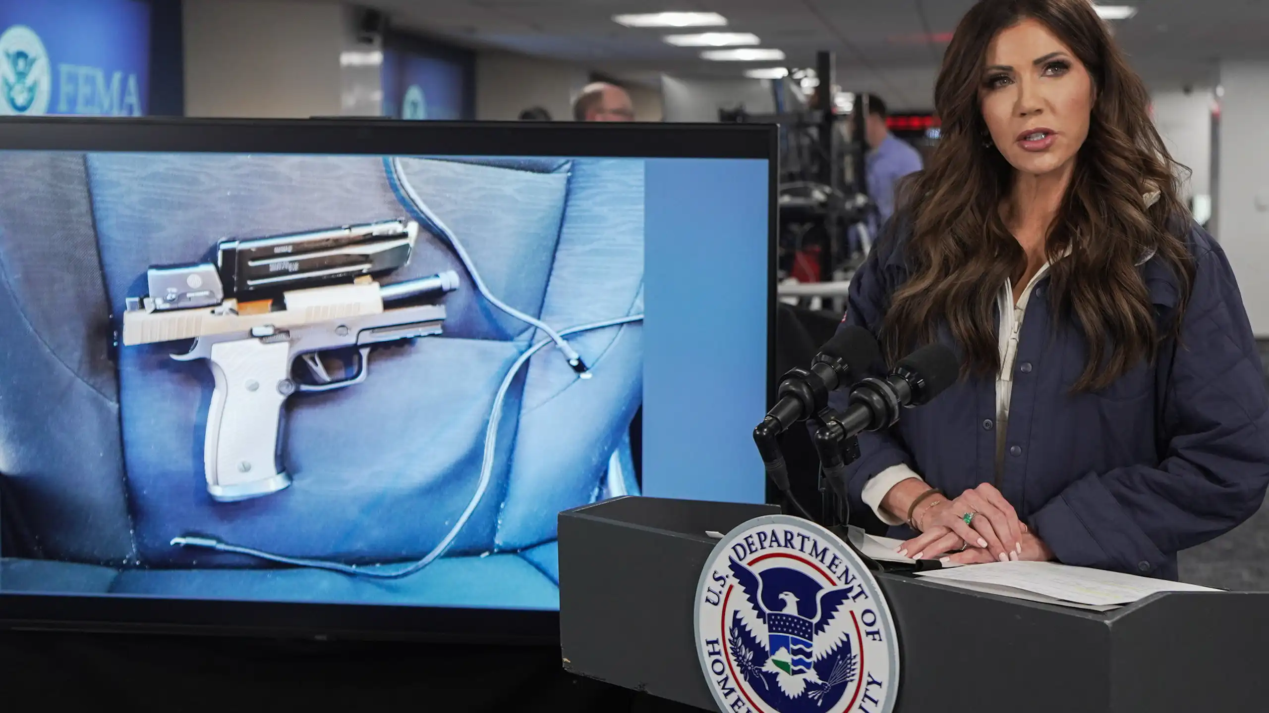 A woman speaks in front of a podium as an image of a gun appears on a video screen behind her.