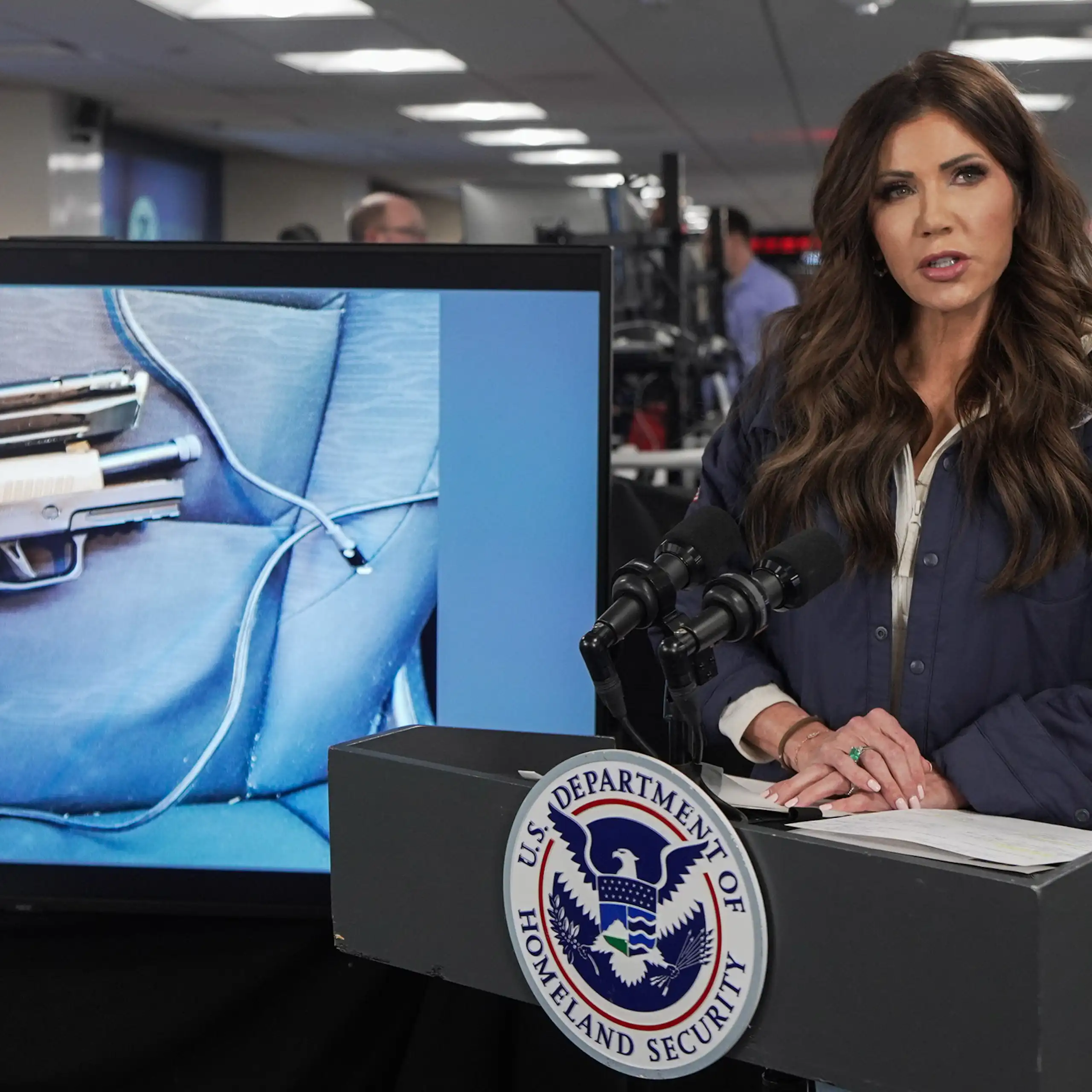 A woman speaks in front of a podium as an image of a gun appears on a video screen behind her.
