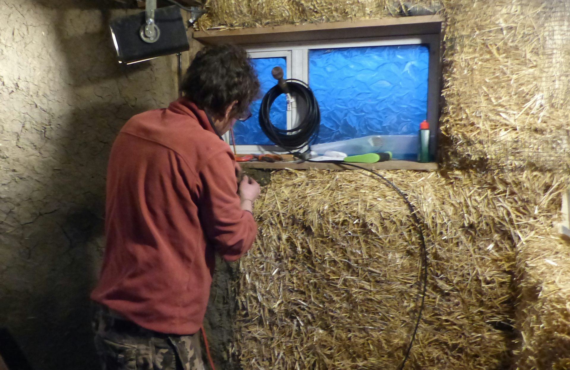 A man wearing red with his hands on a straw bale that is part of a building he is constructing.