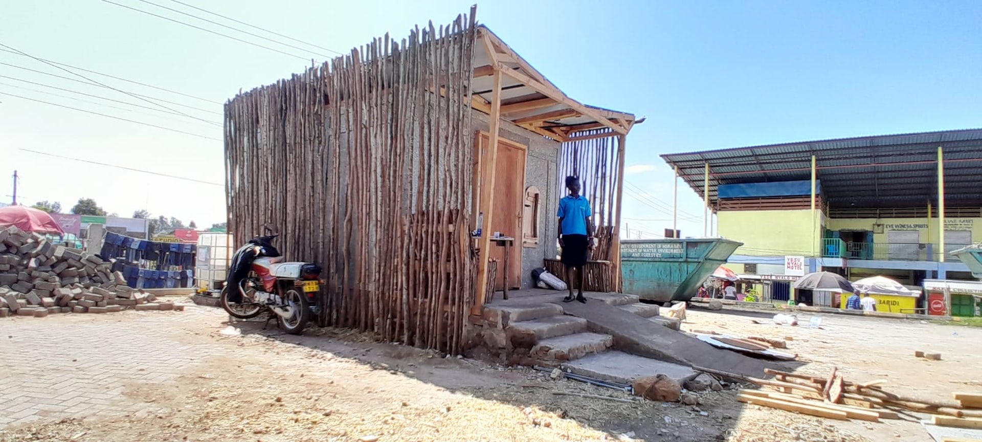 A building at a Kenyan market with a woman stood outside, it was built from straw bales.